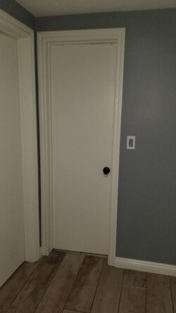 A small, white door with a round black doorknob is framed by light-colored trim, set against a gray wall. On the left, there's another closed white door, and the floor is made of wooden planks. A small, white door with a round black doorknob is framed by light-colored trim, set against a gray wall. On the left, there's another closed white door, and the floor is made of wooden planks.